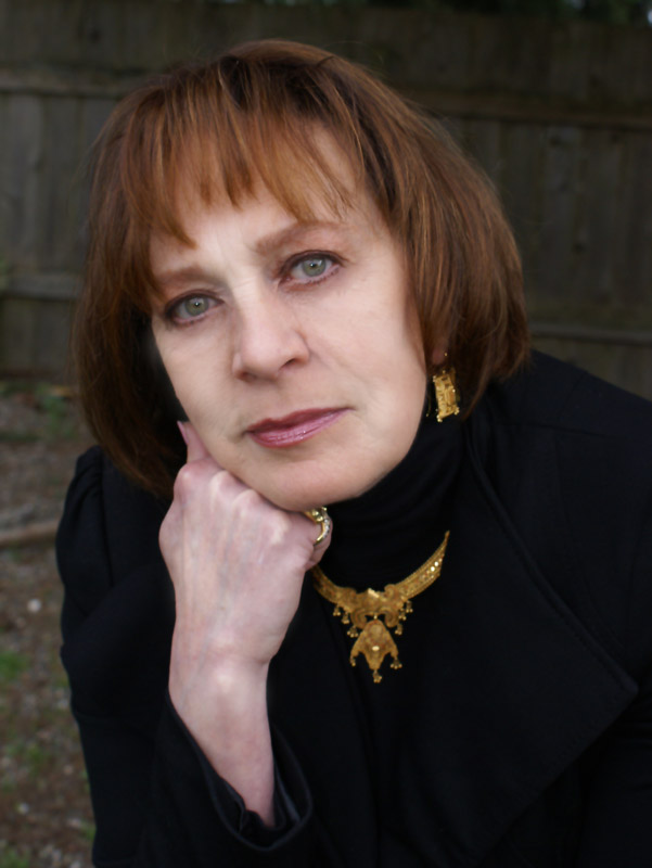 A woman with short brown hair and green eyes poses thoughtfully, wearing a black garment and gold jewelry, including a necklace and earrings. The background features a wooden fence.
